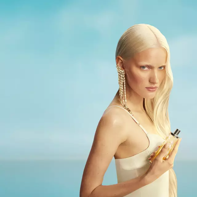 Nouvelle bannière de campagne de Mugler Starlicious Les Eaux Gourmandes mettant en vedette une mannequin aux longs cheveux blonds et aux yeux bleus, vêtue d’une robe jaune pâle et tenant le flacon de la fragrance Pineapple Musk, sur fond de ciel bleu doux. Les parfums sont délicieux, à collectionner et à superposer.