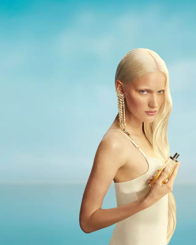 Nouvelle bannière de campagne de Mugler Starlicious Les Eaux Gourmandes mettant en vedette une mannequin aux longs cheveux blonds et aux yeux bleus, vêtue d’une robe jaune pâle et tenant le flacon de la fragrance Pineapple Musk, sur fond de ciel bleu doux. Les parfums sont délicieux, à collectionner et à superposer.